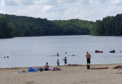 A sandy beach by a lake with people swimming, playing, and relaxing under a partly cloudy sky.