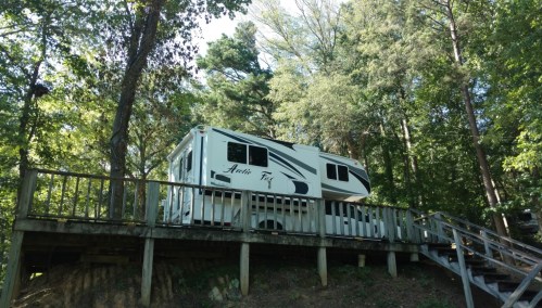 A white RV parked on a wooden deck surrounded by tall trees in a lush green forest.
