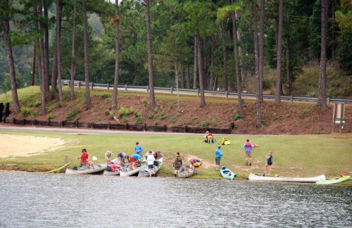 People preparing kayaks and canoes by a lake, surrounded by trees and grassy areas.