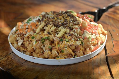 A large platter of loaded tater tots topped with cheese, meats, tomatoes, and green onions on a wooden table.