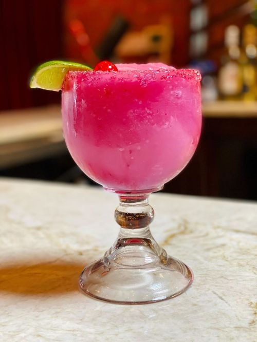 A vibrant pink cocktail in a glass, garnished with a lime wedge and a cherry, sitting on a marble countertop.