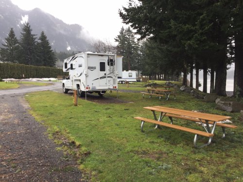 A camper parked near picnic tables in a grassy area, surrounded by trees and mountains in a misty atmosphere.