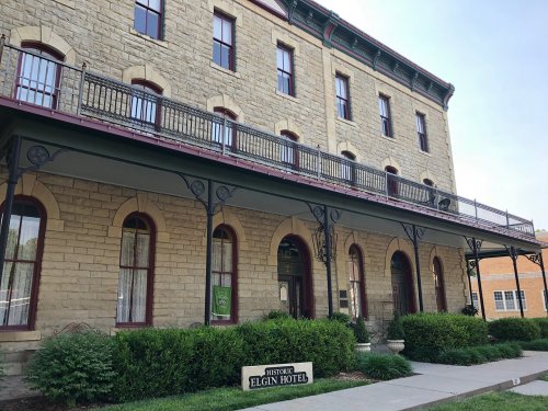 Historic Elgin Hotel, a stone building with a balcony, surrounded by greenery and a clear blue sky.