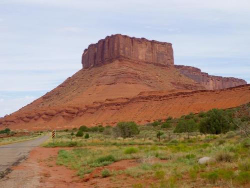 A red rock formation rises sharply from the landscape, surrounded by green shrubs and a winding road in the foreground.