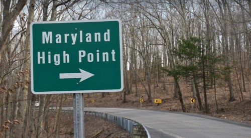 Green road sign indicating "Maryland High Point" with an arrow pointing right, surrounded by bare trees and a winding road.
