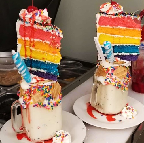 Two colorful milkshakes topped with rainbow cake slices, whipped cream, sprinkles, and a candy cane.