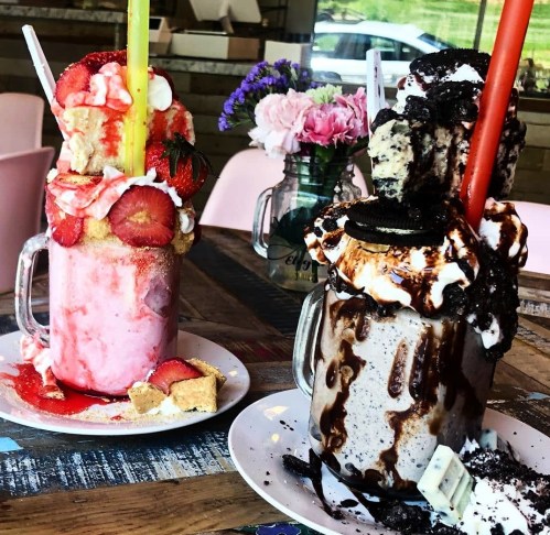 Two extravagant milkshakes topped with whipped cream, cookies, and strawberries, served on colorful plates.