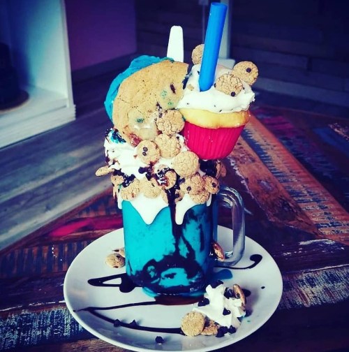 A colorful milkshake topped with whipped cream, a cupcake, cookies, and chocolate syrup, served in a glass mug.