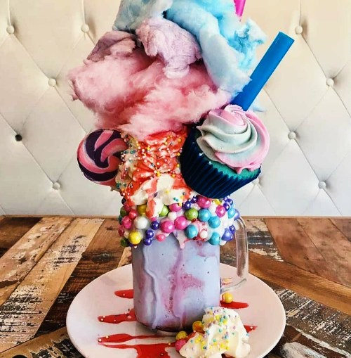 A colorful milkshake topped with cotton candy, cupcakes, sprinkles, and candy, served in a glass on a plate.