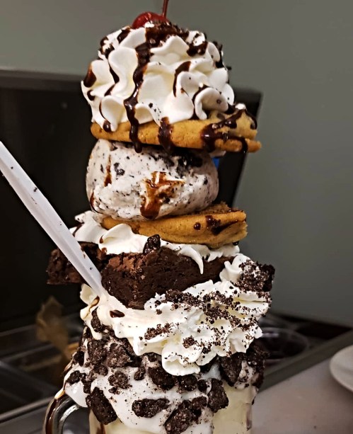 A towering dessert with ice cream, cookies, brownies, whipped cream, chocolate drizzle, and a cherry on top.