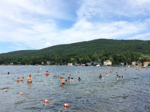 A crowded lake scene with people swimming and enjoying the water, surrounded by green hills and houses in the background.