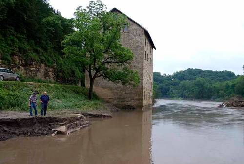 Two people stand near a flooded riverbank, with a stone building partially submerged in the water.