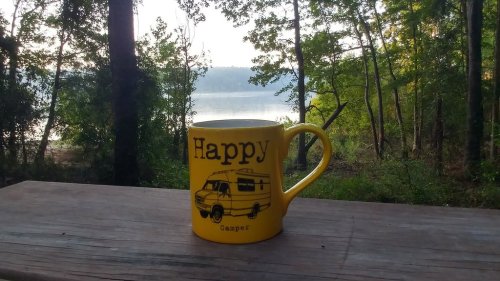 A yellow mug with "Happy Camper" text sits on a wooden table, overlooking a serene lake and trees in the background.