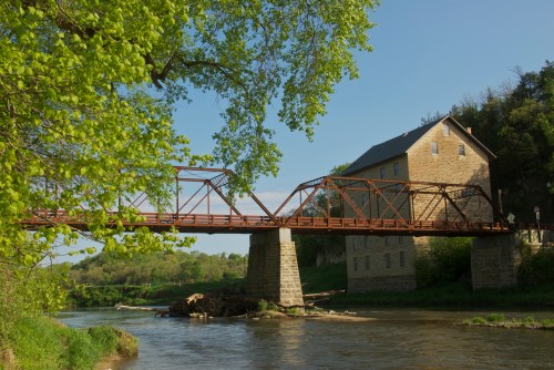 A rustic metal bridge spans a river, with a stone building and lush green trees in the background under a clear blue sky.