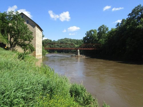 A river flows beside a stone building and a red metal bridge, surrounded by lush green trees and a blue sky.