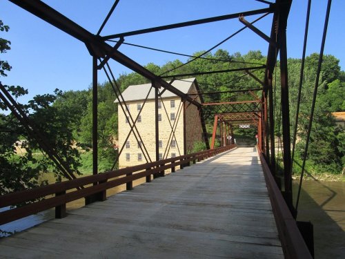 A wooden bridge with metal supports spans a river, leading to a stone building surrounded by trees. Clear blue sky above.