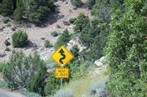Winding road sign indicating curves ahead for the next 2 miles, surrounded by greenery and rocky terrain.