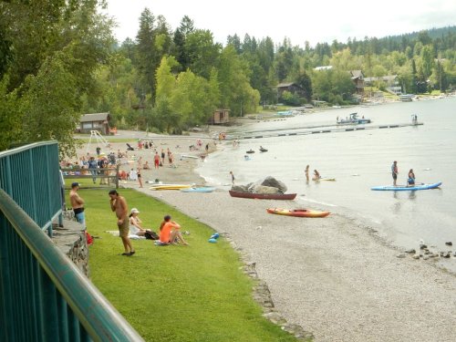A beach scene with people sunbathing, swimming, and kayaking near a lake, surrounded by trees and cabins.