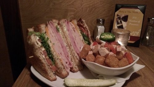 A hearty sandwich with layers of ham, turkey, lettuce, and tomato, served with tater tots and a pickle on the side.