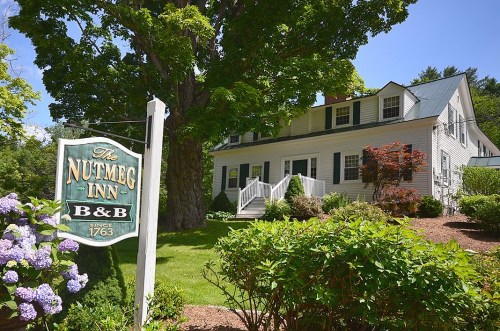 A charming bed and breakfast, The Nutmeg Inn, surrounded by greenery and flowers, with a welcoming sign in front.