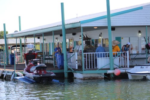 A waterfront building with people, a jet ski, and boats docked nearby on a sunny day.