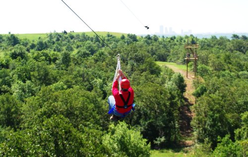 A person in a red helmet and harness zips down a zip line over a lush green landscape with distant city skyline.