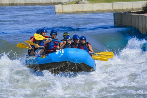 A group of people in a blue raft navigating whitewater rapids, wearing helmets and paddling with yellow oars.