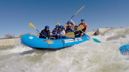 A group of six people in a blue raft navigating whitewater rapids, all wearing helmets and life jackets.