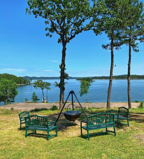 A scenic view of a lake with green benches arranged around a fire pit under clear blue skies and trees.
