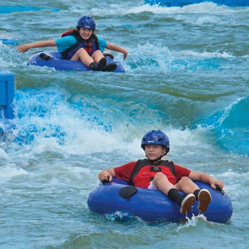 Two children in blue inner tubes ride on a water slide, wearing helmets and enjoying the splashing waves.
