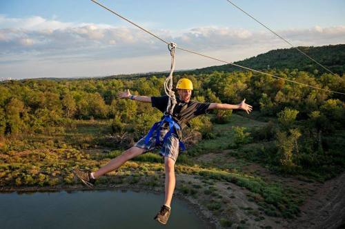 A boy in a yellow helmet zip-lining over a green landscape, arms outstretched and smiling against a blue sky.