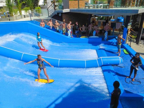 A water park scene with people surfing on a flow rider, surrounded by spectators enjoying the sunny day.