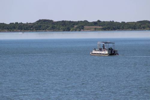A small boat with passengers glides across a calm blue lake, surrounded by green trees in the distance.