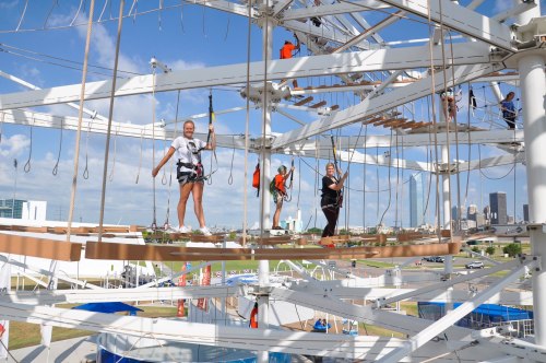 A group of people navigating a high ropes course against a city skyline backdrop on a sunny day.