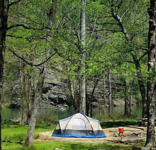 A tent set up in a lush green campsite by a calm river, surrounded by tall trees and rocky cliffs.