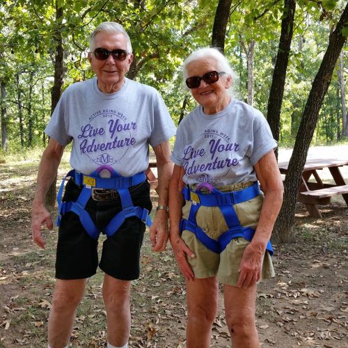 Two elderly individuals wearing matching "Live Your Adventure" t-shirts and harnesses, smiling outdoors in a park.