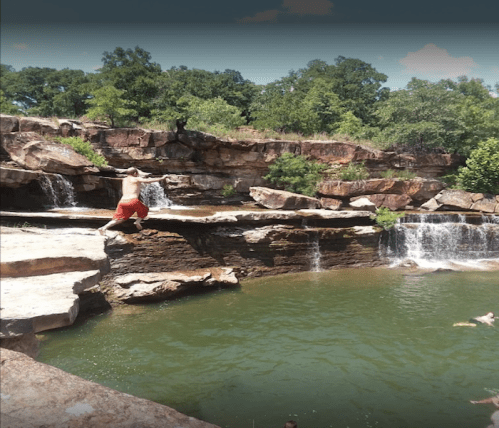 A person jumps off a rock into a green swimming hole surrounded by cliffs and trees. Waterfalls cascade nearby.