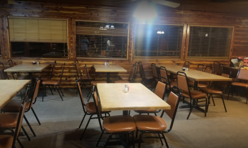 Empty restaurant interior with wooden walls, several tables and chairs, and large windows with blinds.