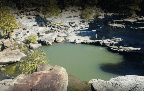 A serene natural pool surrounded by rocky terrain and trees, with people exploring the area.