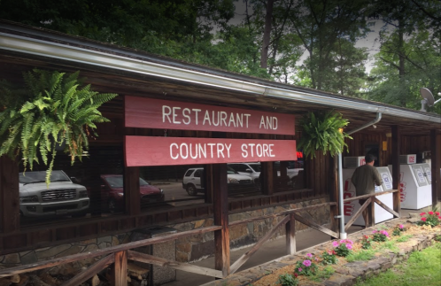 A rustic restaurant and country store with greenery, featuring a wooden sign and a person walking nearby.