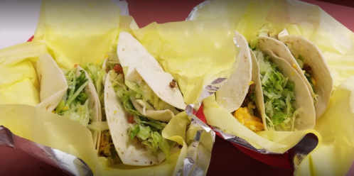 Three tacos in yellow wrappers, filled with lettuce, tomatoes, and meat, served in red baskets.