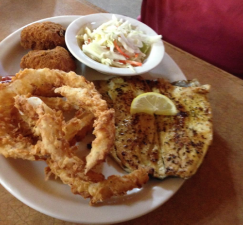 A plate of fried fish with a lemon slice, onion rings, hushpuppies, and coleslaw on the side.