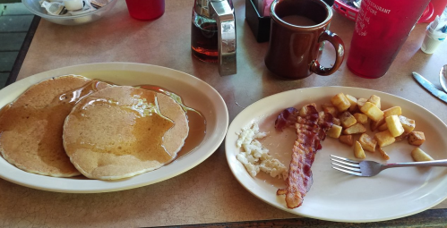 A plate with three pancakes drizzled in syrup, alongside bacon, hash browns, and a cup of coffee.