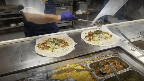 A person in gloves prepares two large burritos with various toppings at a food counter.