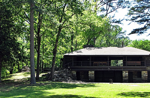 A rustic log cabin surrounded by lush green trees and grass, featuring a stone foundation and a spacious porch.