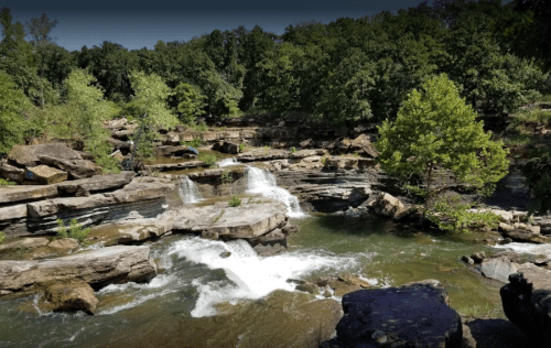 A serene river flows over rocky terrain, surrounded by lush green trees and a peaceful natural landscape.