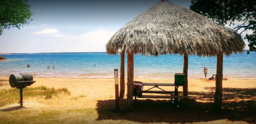 A beach scene featuring a thatched gazebo, picnic table, grill, and people enjoying the water in the background.
