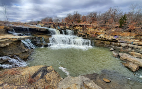 A scenic waterfall cascading over rocky ledges into a calm pool, surrounded by trees and cloudy skies.