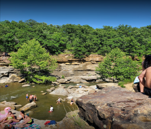 People swimming and relaxing by a rocky river surrounded by lush green trees on a sunny day.