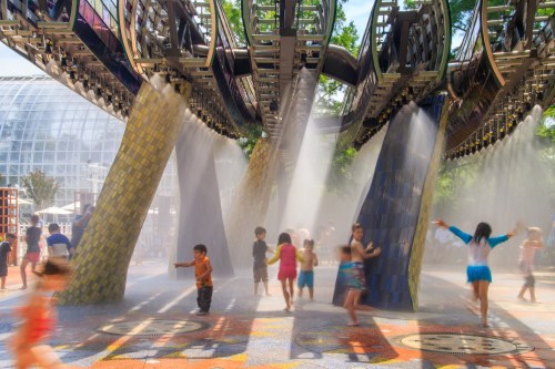 Children play in a splash pad with water jets, surrounded by colorful structures and mist on a sunny day.